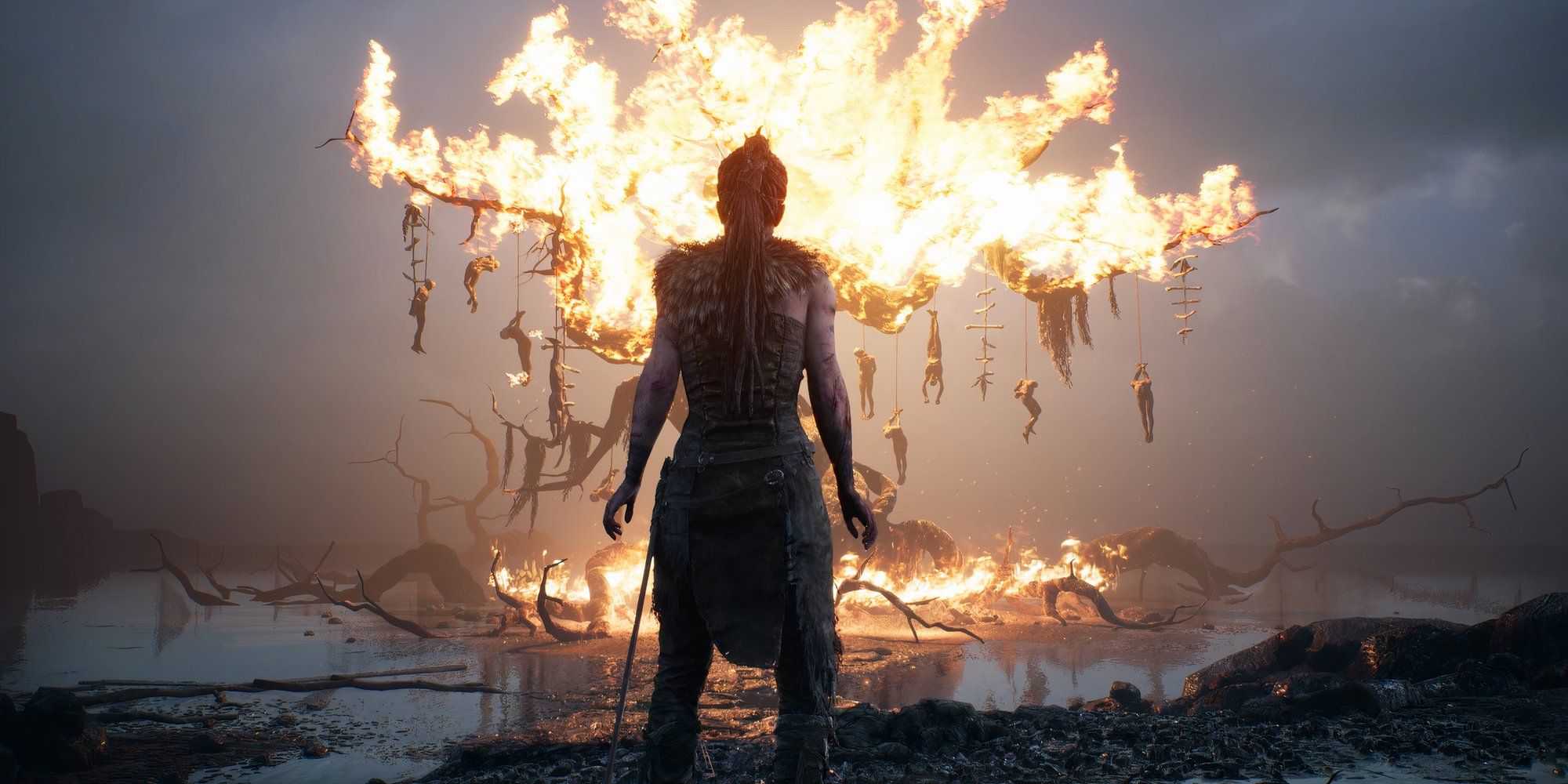 Senua stands in front of a burning tree with hanging bodies in Hellblade Senua's Sacrifice